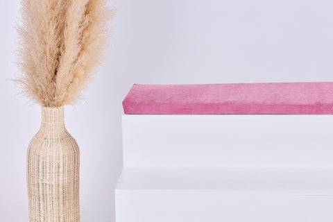 Eine hohe geflochtene Vase mit getrocknetem Pampasgras steht neben einer weißen stufenartigen Struktur, die mit der KraftKids GmbH Sitzauflage für Ikea TROFAST Cord Breitcord Rosa gekrönt ist, vor einem schlichten hellen Hintergrund.