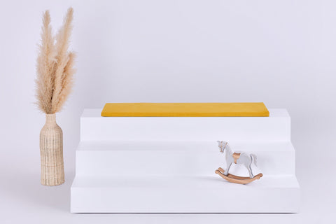 Ein weißer, dreistöckiger Aufsteller mit einer KraftKids GmbH Sitzauflage für Ikea TROFAST Doppelkrepp Gelb Senf auf der oberen Etage, einer dekorativen Vase mit getrocknetem Pampasgras auf der linken Seite und einem kleinen Holzschaukelpferd auf der unteren Etage.