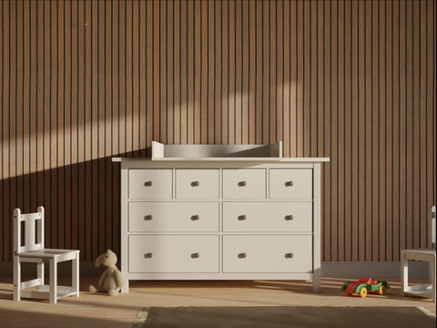 Ein weißer KraftKids Wickelaufsatz (gerade Front) passt auf eine 160 cm lange HEMNES Kommode, die an einer Holzlattenwand steht, neben einem kleinen weißen Stuhl, einem Plüschbären und einem Spielzeugauto auf dem Boden in diesem sanft beleuchteten Raum.