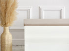 Eine weiße Bank mit der KraftKids GmbH Sitzauflage für Ikea TROFAST Musselin beige steht neben einer geflochtenen Vase mit getrocknetem Pampasgras in einem minimalistischen, modernen Raum mit weiß getäfelten Wänden.