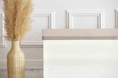 Eine weiße Bank mit der KraftKids GmbH Sitzauflage für Ikea TROFAST Musselin beige steht neben einer geflochtenen Vase mit getrocknetem Pampasgras in einem minimalistischen, modernen Raum mit weiß getäfelten Wänden.