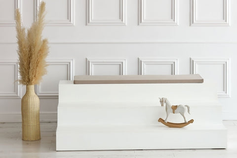 Eine minimalistische weiße Bank mit der KraftKids GmbH Sitzauflage für Ikea STUVA/SMÅSTAD Musselin beige steht vor einer getäfelten Wand. In der Nähe steht eine Weidenvase mit getrocknetem Pampasgras, und auf der Bank ruht ein kleines Holzschaukelpferd.