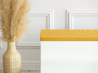Eine hohe geflochtene Vase mit Pampasgras steht neben einer weißen Bank mit der KraftKids GmbH Sitzauflage für Ikea TROFAST Leinen in senfgelb, die an einer weiß getäfelten Wand steht.