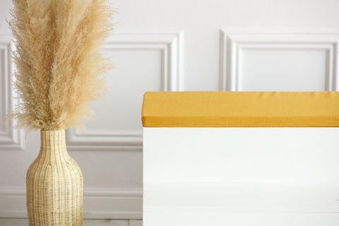 Eine hohe geflochtene Vase mit Pampasgras steht neben einer weißen Bank mit der KraftKids GmbH Sitzauflage für Ikea KALLAX Regal in Leinen gelb senfgelb vor einer weißen Wand mit Zierleiste.