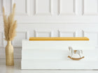 Ein minimalistischer Raum mit einer weiß getäfelten Wand, einer hohen Vase mit Pampasgras, weißen Sitzstufen mit einer KraftKids GmbH Sitzauflage für Ikea TROFAST Leinen in gelb senfgelb und einem kleinen Holzschaukelpferd auf der untersten Stufe.