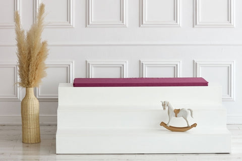 Ein minimalistisches Interieur mit einer hohen Vase mit Pampasgras, einer weißen Stufenbank mit der KraftKids GmbH Sitzauflage für Ikea TROFAST Leinen magenta dunkel und einem kleinen Holzschaukelpferd vor einer weiß getäfelten Wand.