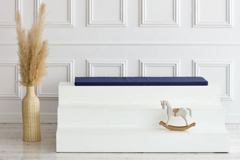 Ein minimalistisches Zimmer mit einer großen beigen Vase mit Pampasgras, einem weißen dreistufigen Podest mit dem KraftKids GmbH Sitzauflage für Ikea TROFAST Leinen blau dunkelblau Kissen und einem kleinen weißen Schaukelpferd. Im Hintergrund eine weiß getäfelte Wand.