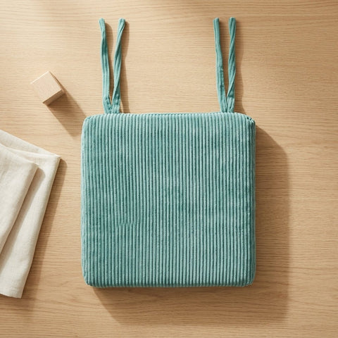 Ein quadratisches, tealfarbenes KraftKids Sitzkissen für Stühle aus Breitcord mit zwei Stoffbändern ruht auf hellem Holz, begleitet von einem beigen Tuch und einem kleinen Holzblock - eine einladende Darstellung des modernen Stils und des Komforts des Kissens.