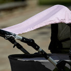 Nahaufnahme eines grauen Kinderwagens mit dem KraftKids Sonnensegel kleine Blätter rosa auf Weiß, das mit weißen Schnüren an den Griffen befestigt ist und den Kinderwagen an einem sonnigen Tag vor einem unscharfen Hintergrund im Freien schützt.