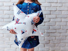 Ein Kind in einem Jeanskleid steht vor einer weißen Backsteinwand und hält das KraftKids Sternkissen Strandhäuschen in der Hand - das perfekte maritime Accessoire für jedes Kinderzimmer.