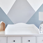 Ein KraftKids Wickelaufsatz Universal weiß mit durchgehendem Boden, eine weiße Kommode mit Schubladen, eine weiße Lampe, ein Kuschelhase, Spielzeug-Schafe in Holzregalen und eine grau-weiße geometrische Akzentwand im Hintergrund.