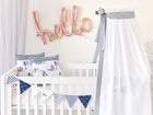 Ein weißes Babybett mit maritimer Bettwäsche aus 100 % Baumwolle und Wimpeln steht neben einem KraftKids Betthimmel dünne Streifen dunkelblau. Roségoldene Luftballonbuchstaben bedeuten "Hallo" und verleihen dem Raum eine leichte und luftige Atmosphäre.