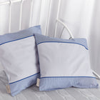 Zwei quadratische Kopfkissen mit KraftKids Kissenbezug Unigrau, mit dünnen dunkelblauen Streifen, liegen auf einer hellgrauen Strukturdecke auf einem weißen Metallbettgestell, mit weißen Vorhängen im Hintergrund.