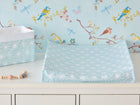 Ein Bezug für Keilwickelauflage weiße Diamante auf Pastell Blau von KraftKids aus 100% Baumwolle liegt auf einer weißen Kommode neben einem passenden Korb, umgeben von bunten Vögeln, Schmetterlingen und Zweigen sowie einer Schnullerkette.