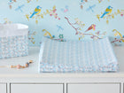 Ein KraftKids-Wickeltisch mit dem "Bezug für Keilwickelauflage kleine Dreiecke blau grau weiß", einer geometrischen blau-grau-weißen Wickelauflage, einem Aufbewahrungskorb und einer Schnullerklemme, vor einer Tapete mit bunten Vögeln und Libellen.