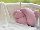 Eine KraftKids Wickeltischumrandung Musselin rosa ist über einen weißen Stuhl drapiert, daneben liegt ein knotenförmiges rosa Kissen mit weißer Paspel vor einem verschwommenen Hintergrund mit Grünzeug.