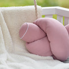 Eine KraftKids Wickeltischumrandung Musselin rosa ist über einen weißen Stuhl drapiert, daneben liegt ein knotenförmiges rosa Kissen mit weißer Paspel vor einem verschwommenen Hintergrund mit Grünzeug.