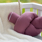 Die KraftKids Wickeltischumrandung Musselin purpur ist auf einer weißen flauschigen Decke auf einer weißen Holzschaukel ausgestellt, mit einem sanften grünen Hintergrund.