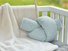 Ein KraftKids Wickeltischumrandung Musselin mint Pusteblumen in Hellgrün mit weißem Pusteblumenmuster liegt auf einer weichen cremefarbenen Decke über einem weißen Kinderbett, akzentuiert durch ein graues Kissen und unscharfe Grünpflanzen im Hintergrund.
