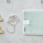 Auf einer hellgrauen Fläche sind kleine tanfarbene Babyschuhe, ein grau-weiß gestreiftes Baby-Bonnet und die KraftKids Reisewickelunterlage Musselin mint Pusteblumen arrangiert.