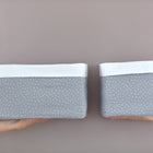 Zwei Hände halten zwei KraftKids Körbchen Musselin grau Punkte - handgenähte Baumwollkörbchen mit weißem Deckel und grauem Tupfenkörper, ideal zur Aufbewahrung auf dem Wickeltisch. Der linke Korb ist größer, beide stehen vor einem hellen Hintergrund.