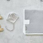 Ein Paar beigefarbene Babyschühchen, eine grau-weiß gestreifte Babymütze und eine gefaltete KraftKids Reisewickelunterlage Musselin grau Punkte sind ordentlich auf einer hellen, strukturierten Oberfläche angeordnet.