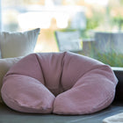 Ein KraftKids qualitäts Stillkissen Waffel Piqué rosa mit ergonomischer Form ruht auf einem dunklen Sofa, umgeben von beigen und weißen Kissen in einem hellen, luftigen Raum.