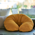 Ein KraftKids qualitäts Stillkissen Waffel Piqué Senf mit ergonomischem Design ruht auf einem dunklen Sofa, umgeben von hellen Kissen und einer cremefarbenen Decke, während im Hintergrund das Sonnenlicht durch das Fenster fällt.