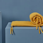 Die KraftKids Bettumrandung Nestchen Waffel Piqué Senf aus 100% Baumwolle ist teilweise gerollt, mit Stoffbändern gebunden und auf einem hellblauen rechteckigen Sockel vor blauem Hintergrund platziert.