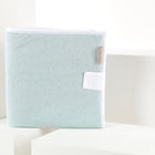 Die Wickelauflage aus grünem Musselin mit goldenen Punkten von KraftKids steht aufrecht auf weißen geometrischen Blöcken und ist mit einem beigen Etikett an der Seite und einer weißen Lasche auf der Vorderseite versehen.