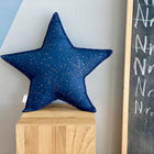 Das KraftKids Sternkissen Musselin mit goldenen Punkten auf Grau ruht auf einem Holzpodest neben einer Tafel. Die modern gestaltete Wand dahinter macht das Kissen zum Highlight für jedes stilvolle Kinderzimmer.