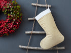 Eine KraftKids Weihnachtssocke mit goldenen unregelmäßigen Punkten auf olivem Grün hängt an einer rustikalen Holzleiter vor grauer Wand. Links ergänzt ein grüner Kranz mit roten Beeren die festliche Kinderzimmerdeko.