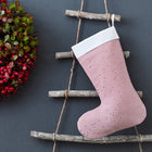 Eine KraftKids Weihnachtssocke Musselin goldene Punkte auf Rosa hängt an einer rustikalen Holzleiter vor grauer Wand. Links davon ergänzt ein festlicher Kranz mit roten Beeren und grünen Blättern die Weihnachtsdekoration.