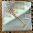 Die Reisewickelunterlage Leinen leicht grünes Weiß von KraftKids liegt zusammengefaltet auf Rattan, darüber ein getrockneter Grashalm und sanftes Sonnenlicht zaubert zarte Schatten auf die Wickelunterlage.