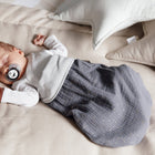 Ein Baby schläft in einem beigen Bett mit cremefarbenen und weißen Sternenkissen. Es trägt ein weißes Hemd und den KraftKids Strampelsack Frühling Sommer Musselin goldene Punkte auf Grau, zusammen mit einem Schnuller mit Bärengesicht.