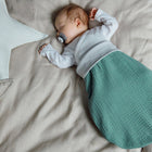 Ein Baby mit einem Schnuller schläft auf einem beigen Bett, trägt ein weißes Oberteil und den KraftKids Strampelsack Herbst Winter Musselin nilblau. Ein großes, weiches, sternförmiges Kissen liegt neben dem Baby.