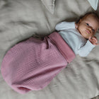 Ein Baby liegt auf einem weichen, hellen Bettchen, trägt ein weißes, langärmeliges Hemdchen und ist kuschelig in den KraftKids Strampelsack Herbst Winter Musselin rosa eingewickelt. Es blickt mit großen Augen auf und hält eine Hand vor das Gesicht.