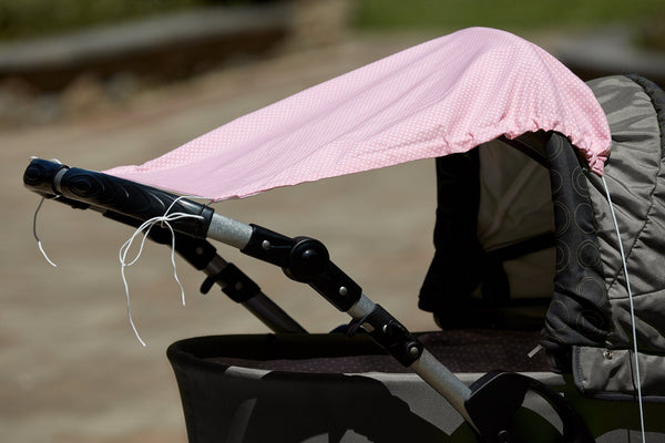 Eine Nahaufnahme eines KraftKids Sonnensegels "weiße Punkte auf Koralrosa" ist mit weißen Schnüren an einem Kinderwagengriff befestigt. Der Baumwollstoff hebt sich ab, während der unscharfe Hintergrund auf den Bürgersteig und das Grün im Freien hindeutet.