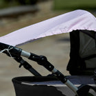 Eine Nahaufnahme eines KraftKids Sonnensegel weiße Punkte auf Rosa zeigt sein hellrosa, gepunktetes Verdeck, das lose an einem grau-schwarzen Kinderwagengestell befestigt ist, wobei leichte Ausfransungen sichtbar sind, vor einem unscharfen Hintergrund im Freien.