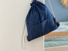 Ein KraftKids Turnbeutel Sternenhimmel in Marineblau mit goldenen Sprenkeln hängt an einer weißen Wand, mit einem Teil eines Holzbettes und hellblauer Bettwäsche im Hintergrund - ein charmantes personalisiertes Geschenk für Kinder.