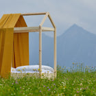 Ein KraftKids Bettbaldachin Musselin gelb Pusteblumen krönt ein minimalistisches Holzbett mit weißer Bettwäsche, das in einem Wildblumenfeld vor nebligen Bergen steht - die perfekte verträumte Kinderzimmer Deko Inspiration.