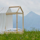 Ein einfaches hölzernes Bettgestell in Form eines Hauses steht in einem grasbewachsenen Feld mit Wildblumen, umhüllt von der KraftKids Bettbaldachin Musselin mit goldenen Punkten auf Weiß. Im Hintergrund erheben sich neblige Berge und ein bewölkter Himmel.