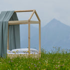Ein minimalistisches Holzbettgestell mit dem Betthimmel von KraftKids Bettbaldachin Musselin mint steht in einer Wildblumenwiese, in der Ferne Berge unter einem wolkenverhangenen Himmel.
