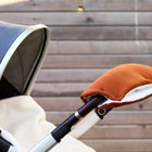 Nahaufnahme eines Kinderwagens mit grauem Verdeck und weißer Decke, an dessen Lenkstange KraftKids Kinderwagenmuff Doppelkrepp Rot Herbstrot Fäustlinge befestigt sind, vor dem Hintergrund eines Holzzauns.