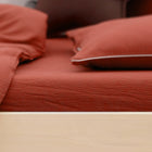 Eine Nahaufnahme eines ordentlich gemachten Bettes mit roter Bettwäsche, auf der das KraftKids Spannbettlaken Doppelkrepp Rot Herbstrot, eine Decke und zwei Kissen auf einem hellen Holzrahmen liegen.