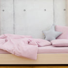 Ein helles Holzbett mit dem KraftKids Bettwäscheset Doppelkrepp Rosa, zwei rosa Kissen und einem grauen Sternkissen sitzt vor einer Betonwand mit runden Einbuchtungen für erholsamen Schlaf.