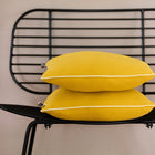 Zwei KraftKids Kissenbezug Doppelkrepp Gelb Senf sind auf einem schwarzen Stuhl mit Drahtgestell vor einer beigen Wand gestapelt.