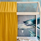 Eine KraftKids Bettbaldachin Doppelkrepp Gelb Senf drapiert sich über ein hölzernes Bett in Hausform vor einem blau-grünen Wandbild mit Walen. Eine kuschelige beigefarbene Decke spendet Wärme und schafft einen stilvollen Musselin-Kinderzimmer-Look.