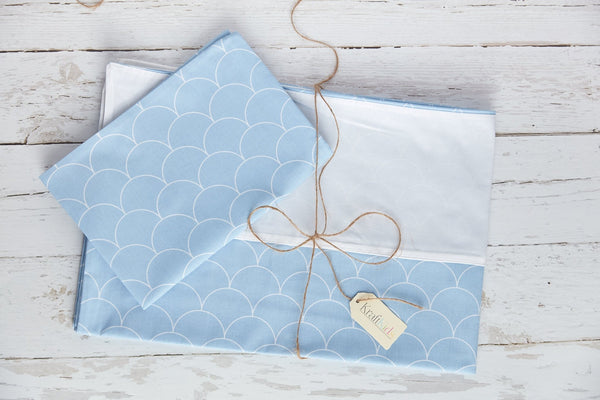 Ein gefaltetes KraftKids Bettwäscheset Uniweiss (weiße Halbkreise auf Pastelblau) aus 100% Baumwolle liegt auf weißem Holz, gebunden mit brauner Schnur und "Kristall" Tag - für erholsamen Schlaf.