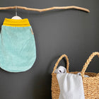 Ein mintfarbener Strampelsack Frühling Sommer Cord Breitcord von KraftKids hängt an einem Holzast über einem geflochtenen Korb mit einer zusammengerollten Decke und einem hellgrauen Tuch an einer dunklen Wand.